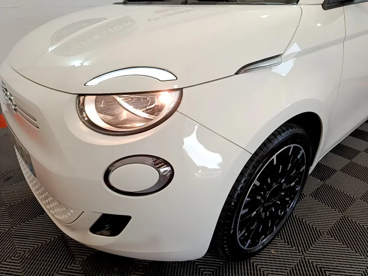 Vue 3/4 avant droit d'une Fiat 500 électrique blanche avec phare allumé et jante noire design moderne.