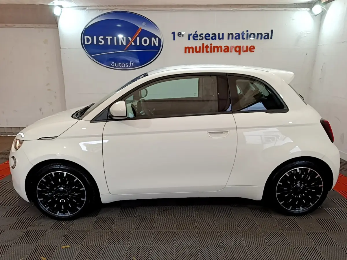 Profil côté gauche d'une FIAT 500 électrique blanche 42kWh Icône 2022 avec jantes noires stylisées en intérieur showroom.
