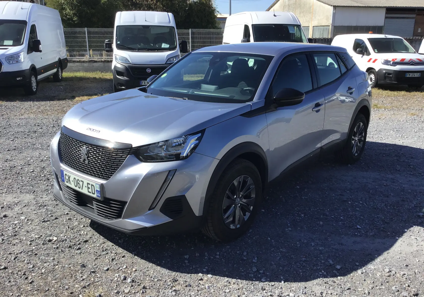 Peugeot 2008 gris Artense en 3/4 avant droit, avec calandre noire et jantes alliage foncé, stationnée sur un sol gravillonné.