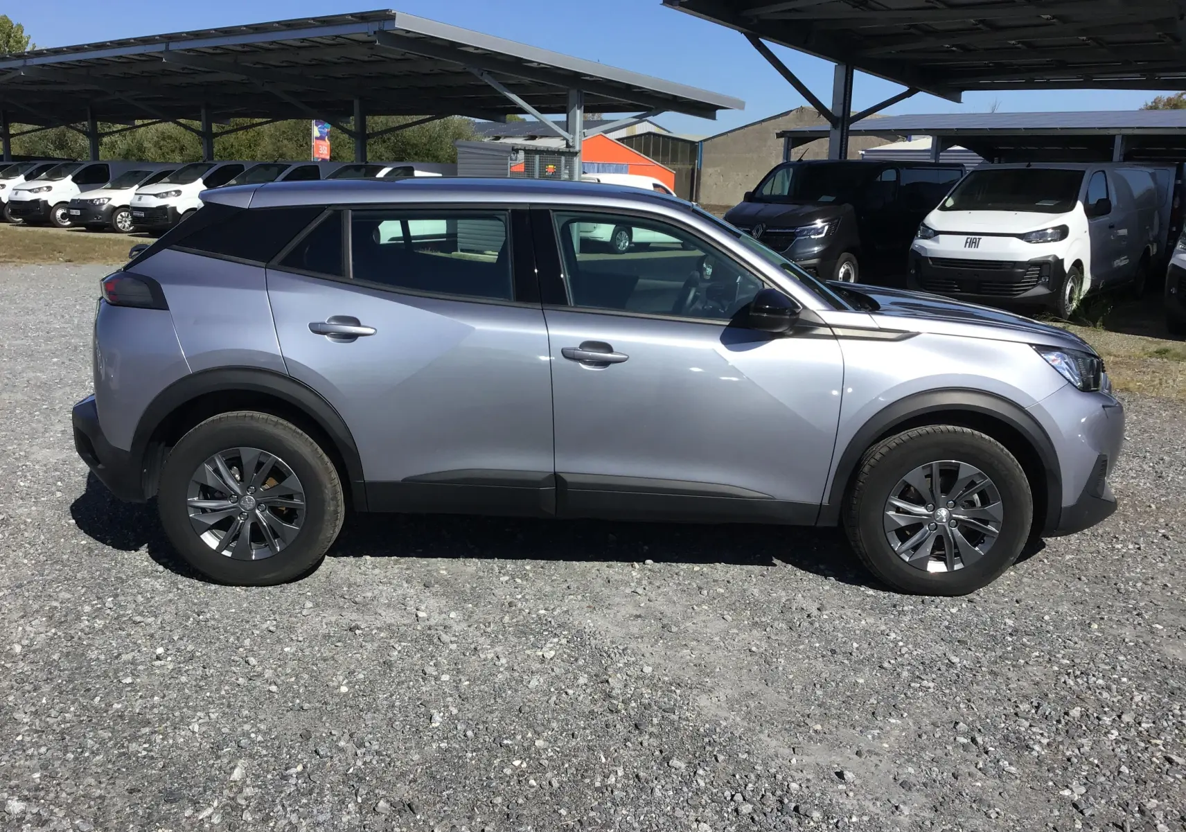 Peugeot 2008 gris Artense vue de profil côté gauche sur parking gravillonné avec protections noires sur les passages de roues.