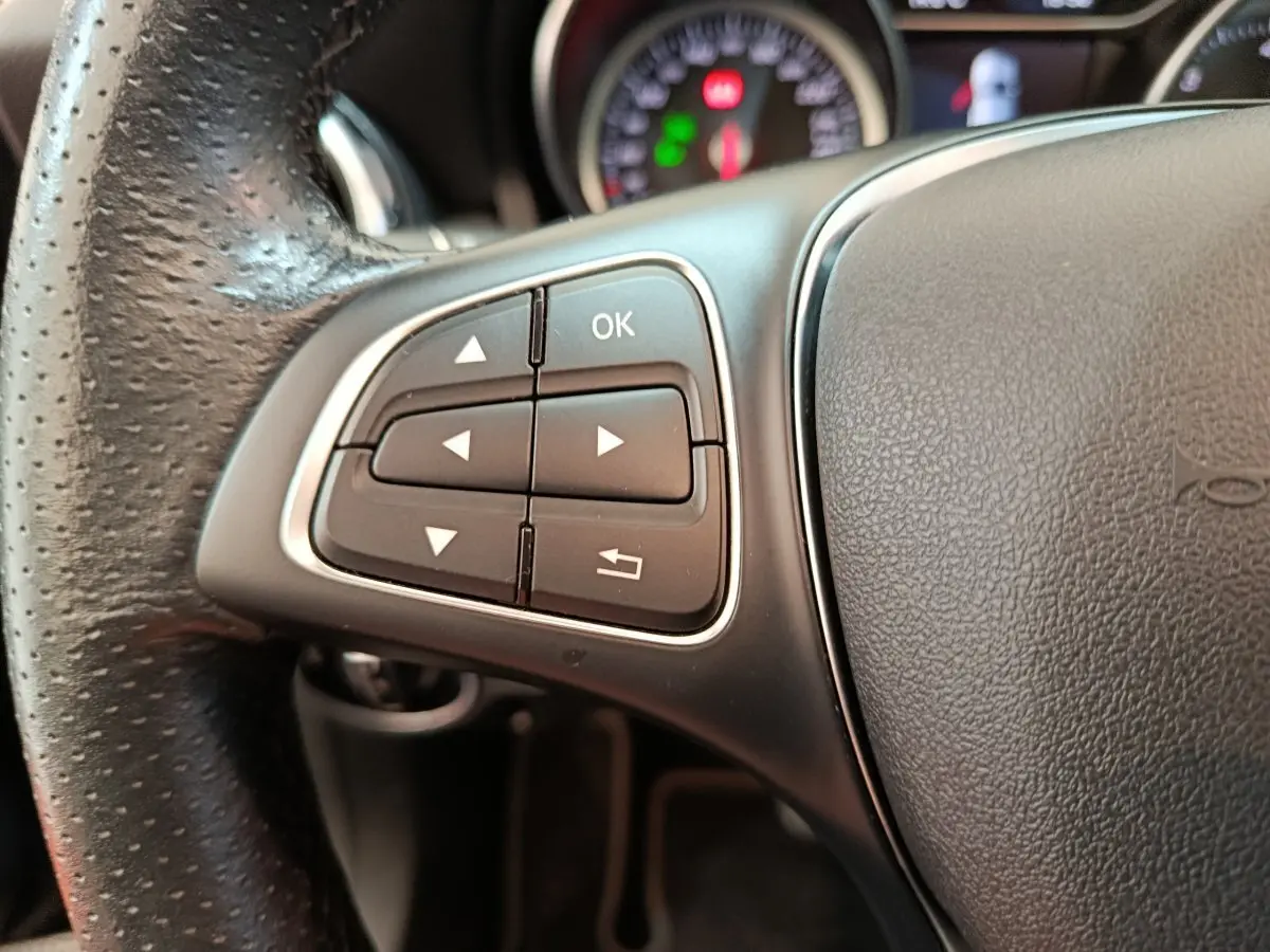 Gros plan sur les commandes gauche du volant en cuir noir du Mercedes GLA 220 d Business Exec 2018, avec tableau de bord flou.