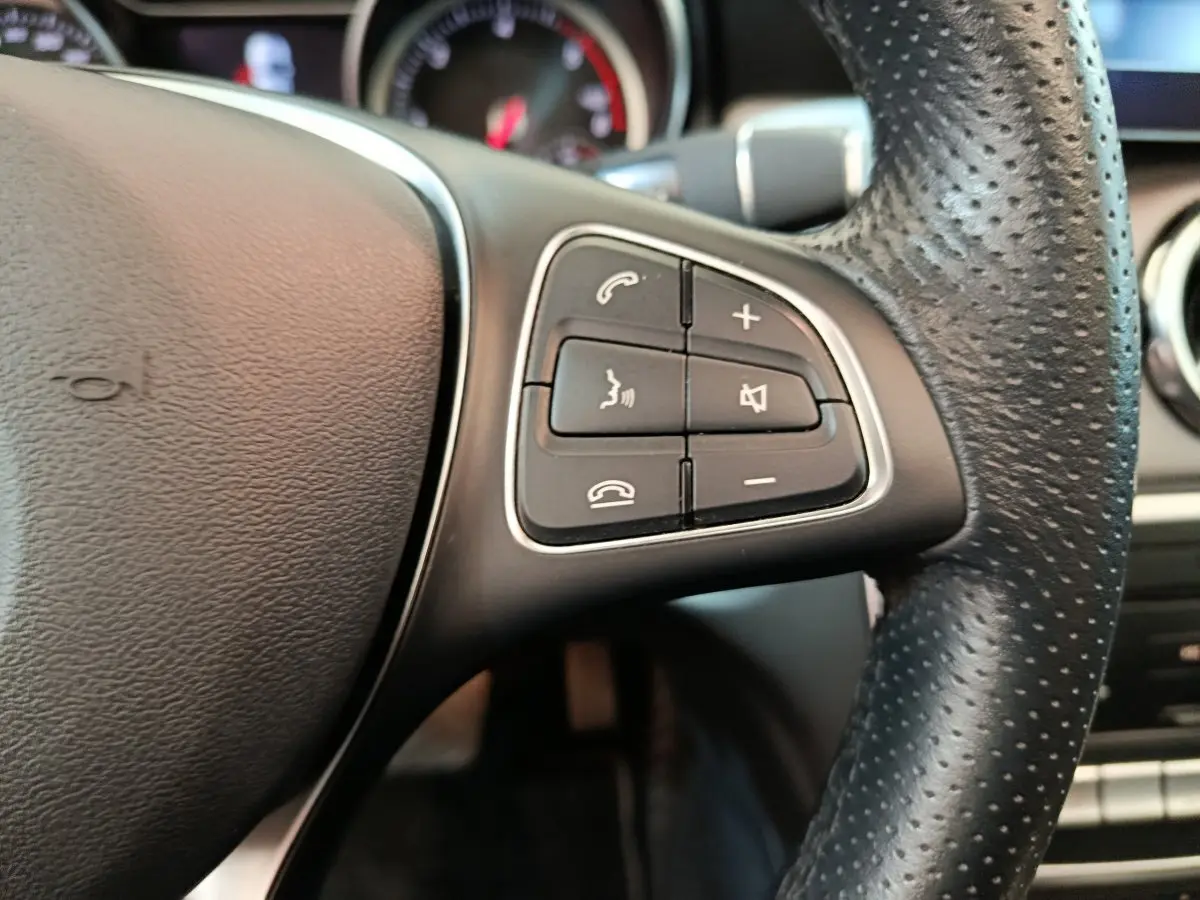 Gros plan sur les commandes audio et téléphone du volant cuir perforé côté gauche d'un Mercedes GLA noir de 2018.