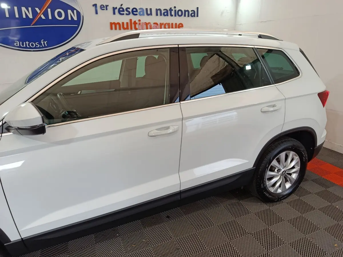 Profil droit d'un Skoda Karoq blanc 2024 avec jantes alliage et barres de toit noires en intérieur showroom.