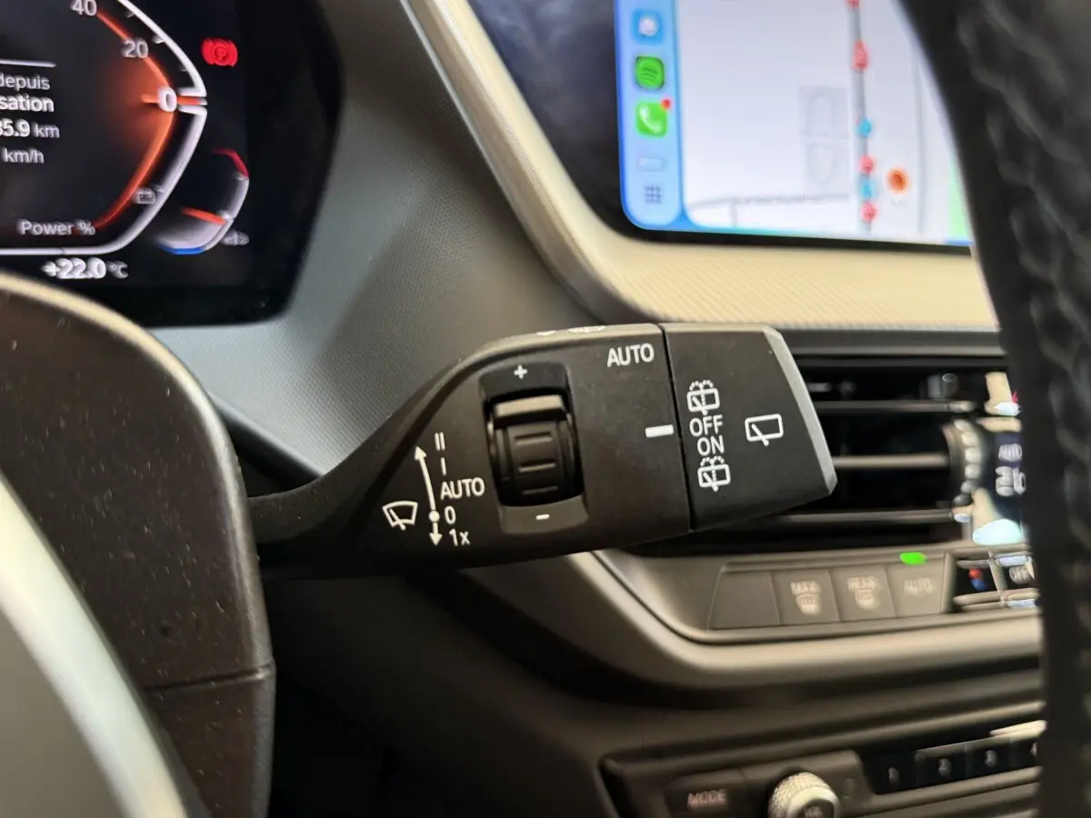 Gros plan sur la commande d’essuie-glace noire dans l’habitacle d’une BMW Série 1 noire, avec tableau de bord et écran tactile visibles.