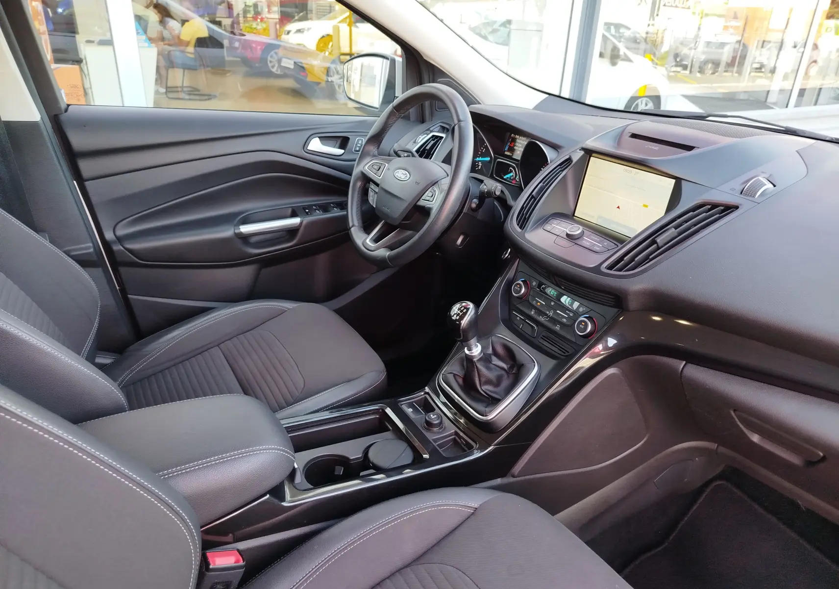 Vue intérieure côté conducteur du Ford Kuga 2019, tableau de bord noir avec écran tactile et levier de vitesses manuel.