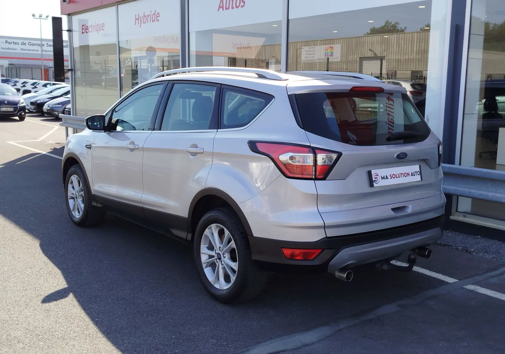 Vue 3/4 arrière droite d'un Ford Kuga gris clair 2019 avec barres de toit et double sortie d'échappement visible.