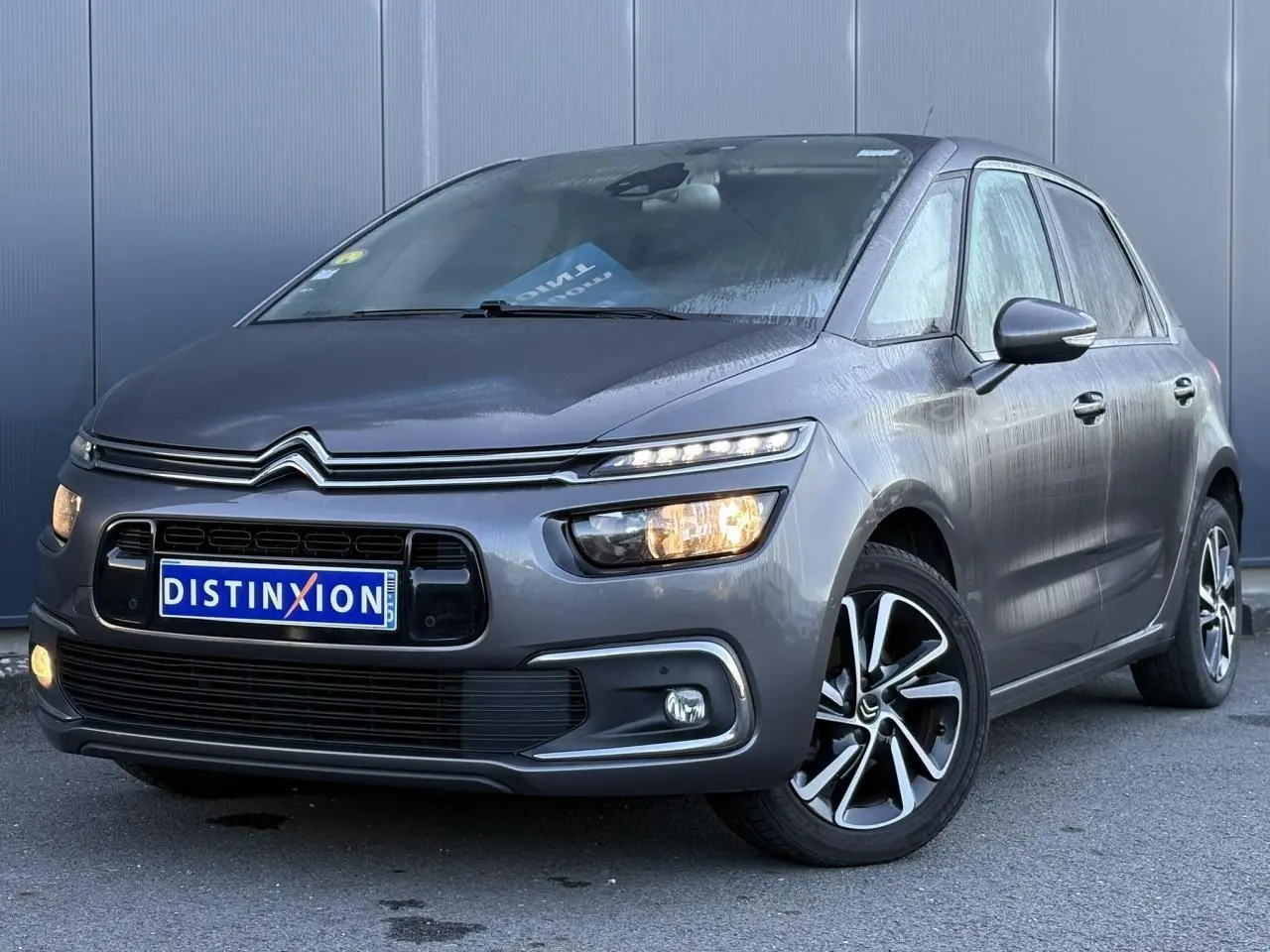 Citroën C4 Picasso gris Platinium vue 3/4 avant droit avec feux allumés et jantes alliage bi-ton.
