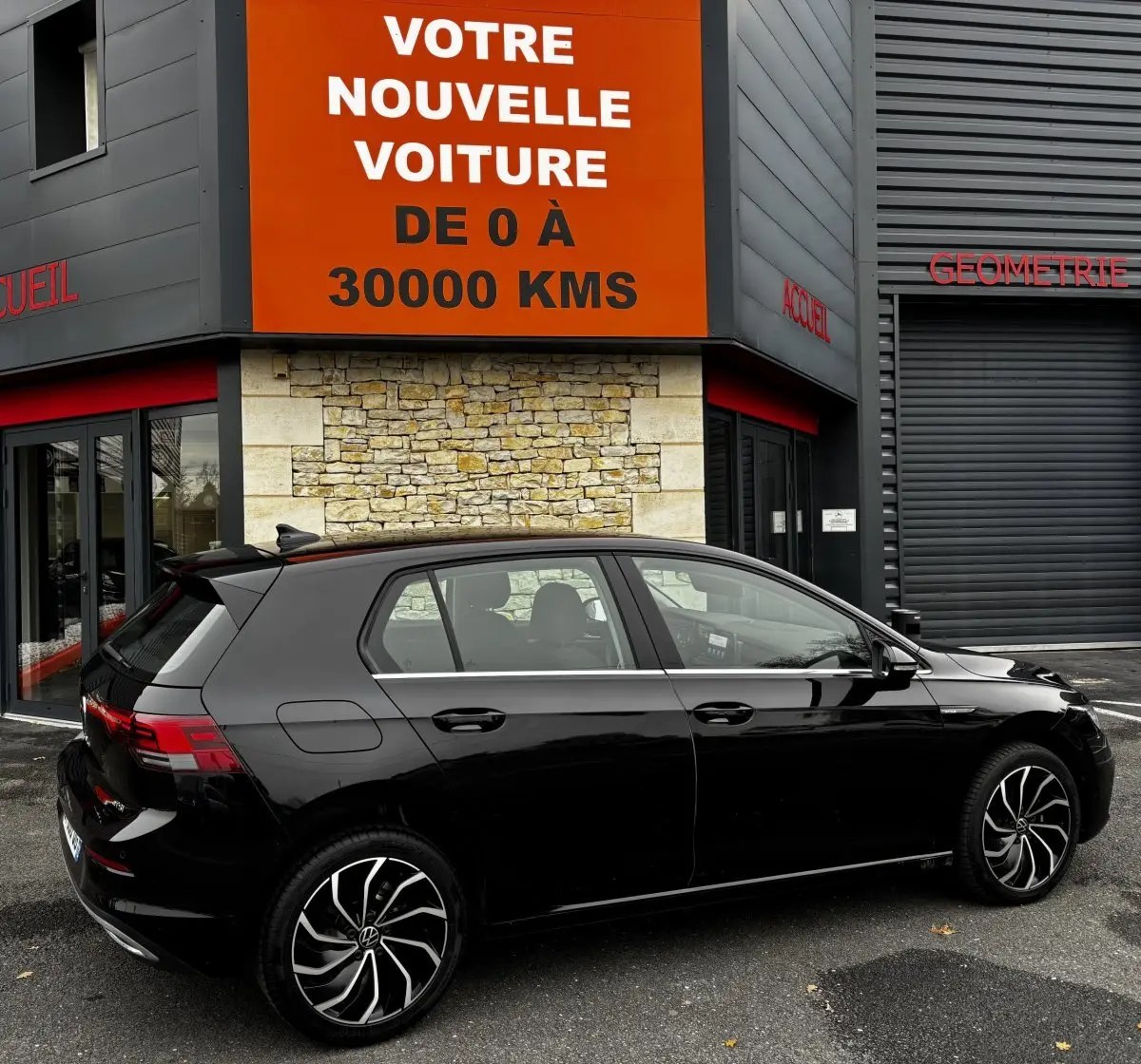 Volkswagen Golf noire vue de profil côté gauche, stationnée devant un bâtiment avec panneau orange "Votre nouvelle voiture".