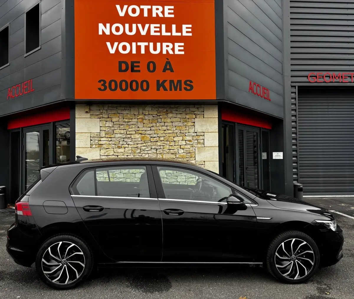Profil côté gauche d'une Volkswagen Golf noire 2024 garée devant un bâtiment avec panneau orange "Votre nouvelle voiture".