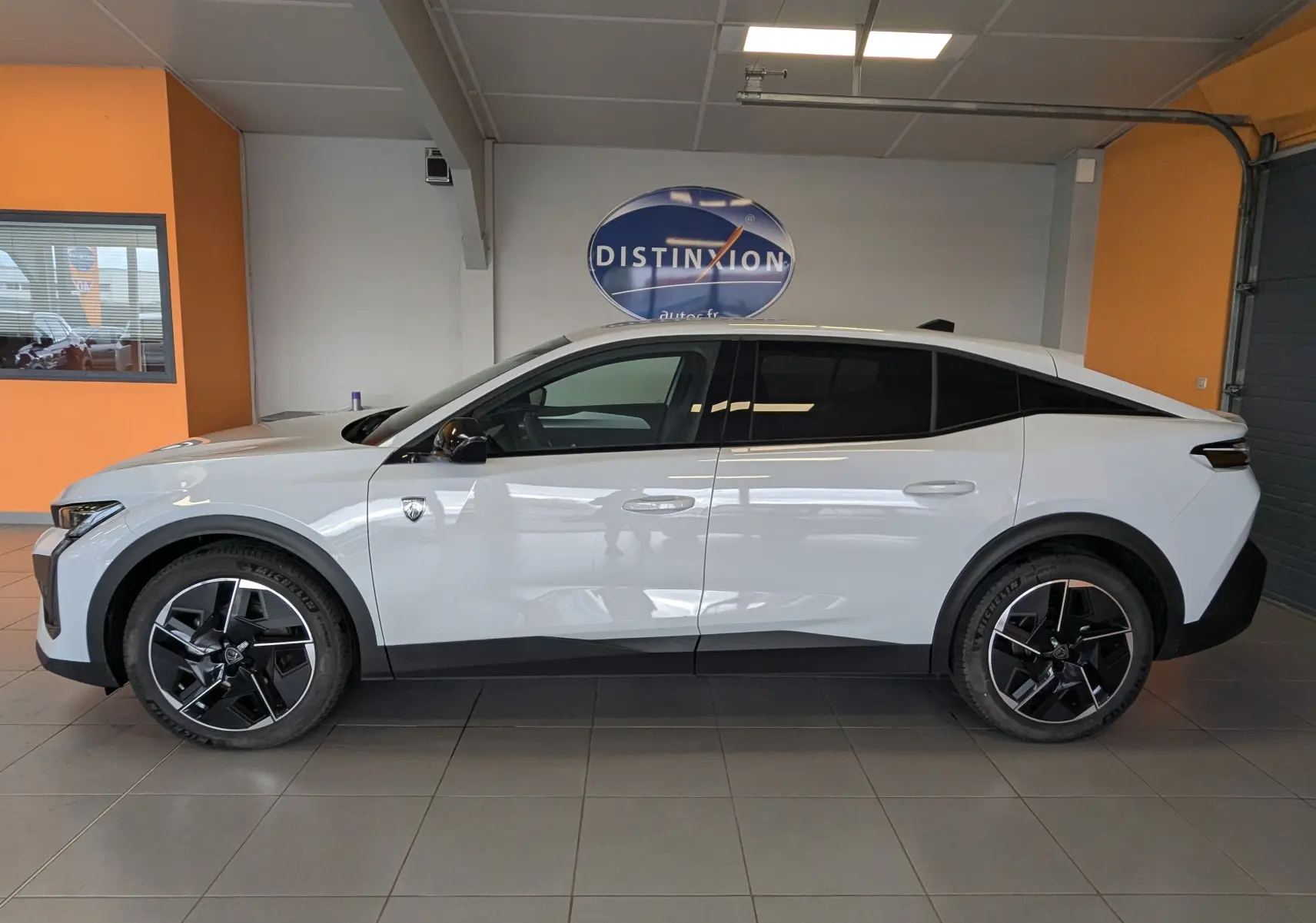 Vue latérale droite d'une Peugeot 408 Hybrid 145 e-DCS6 GT blanche Okenite avec jantes alliage 19 pouces bicolores en intérieur showroom.