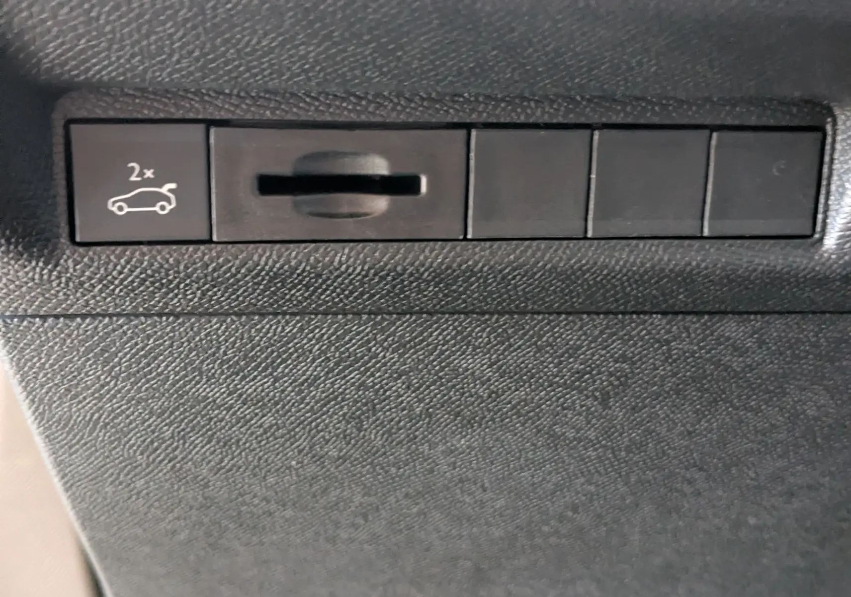 Bouton intérieur noir avec pictogramme d'ouverture du coffre deux fois, sur tableau de bord texturé noir.