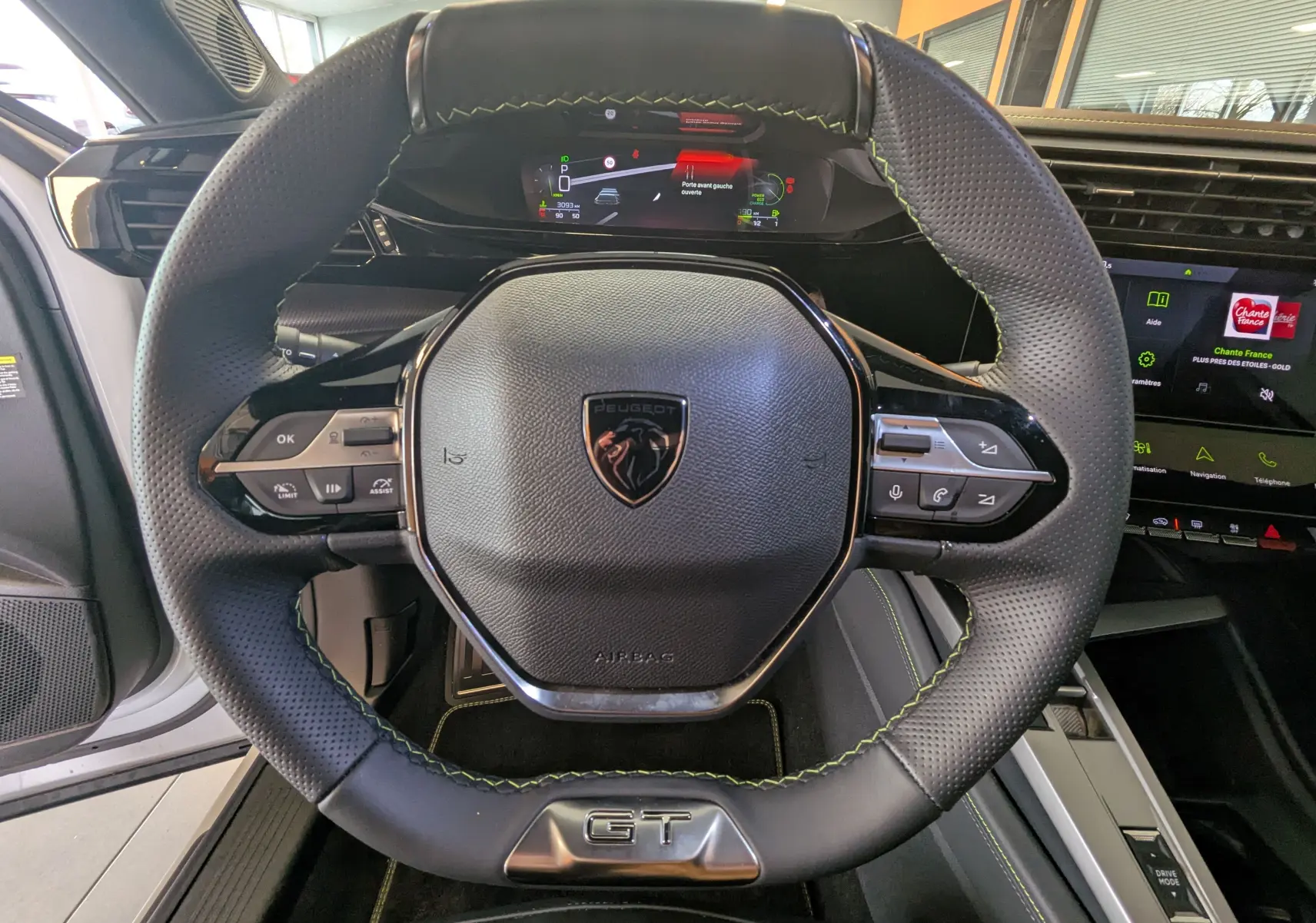 Vue rapprochée du volant GT en cuir noir avec surpiqûres vertes du Peugeot 408 Hybrid 145 e-DCS6, tableau de bord digital visible.
