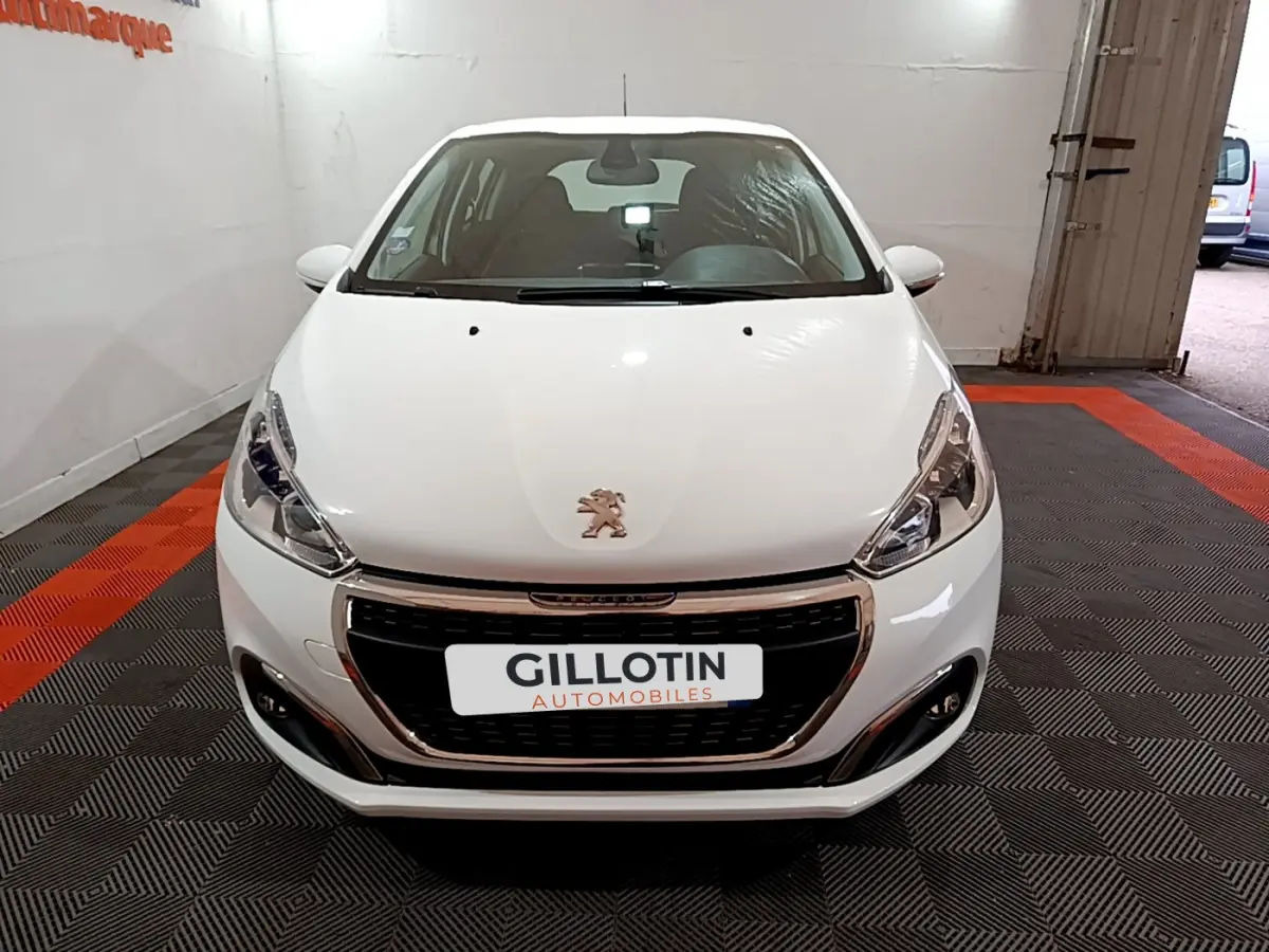 Vue frontale d'une Peugeot 208 Puretech 82 Signature blanche avec calandre chromée et phares LED dans un garage.