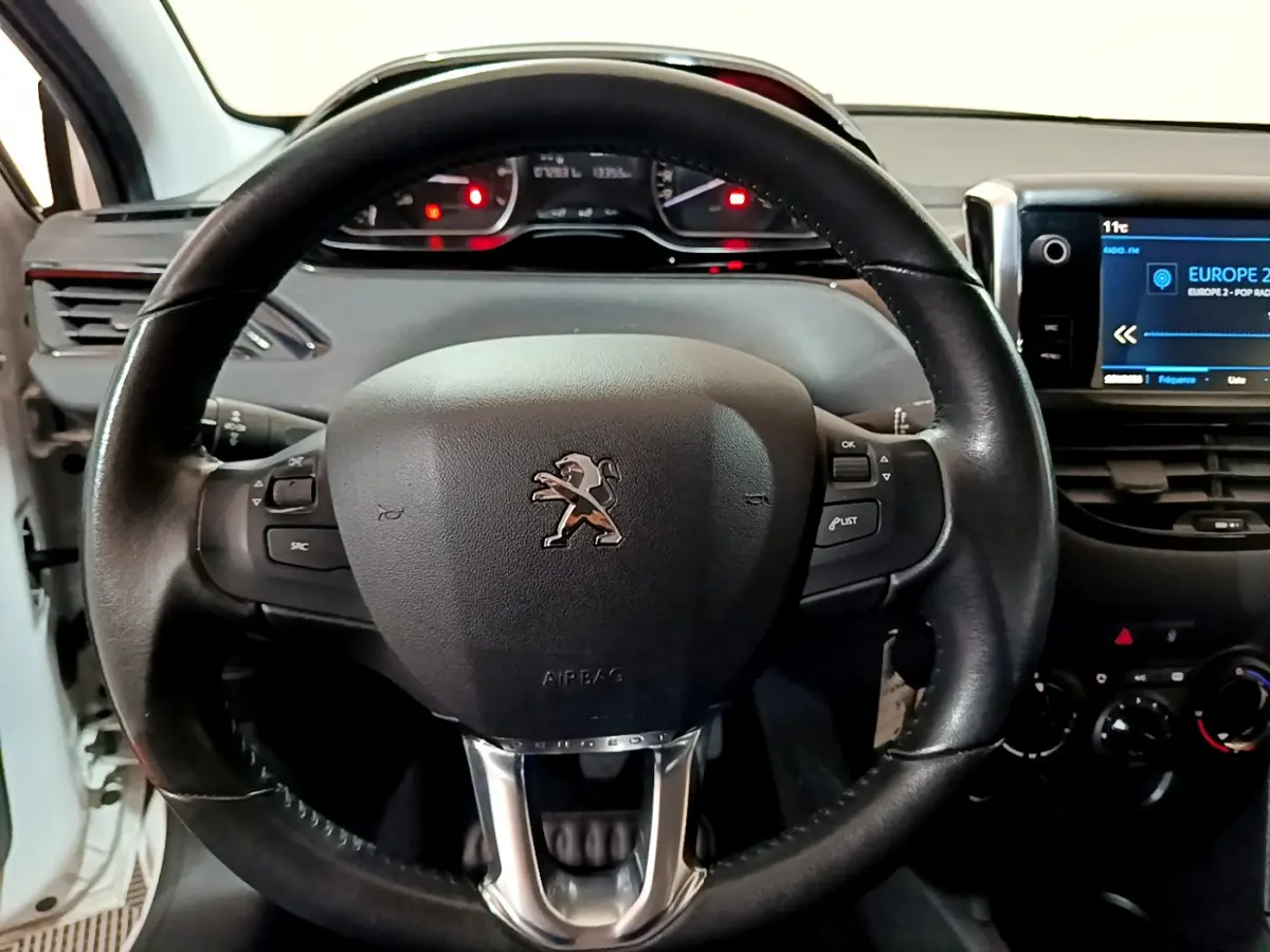 Vue rapprochée du volant cuir noir avec logo Peugeot et tableau de bord numérique d'une 208 blanche de 2018.