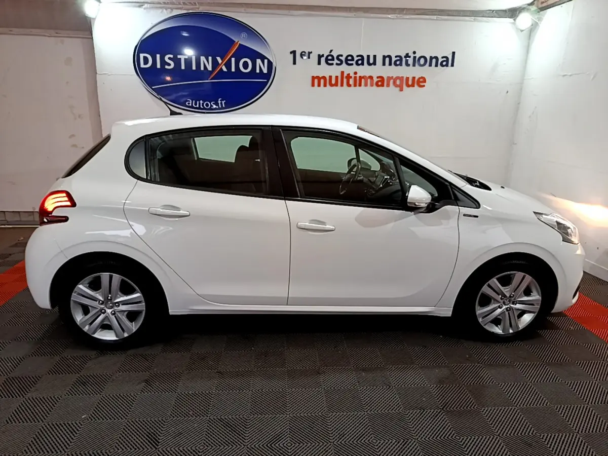 Profil côté gauche d'une Peugeot 208 Puretech 82 Signature blanche, en intérieur sur sol damier avec feux arrière allumés.