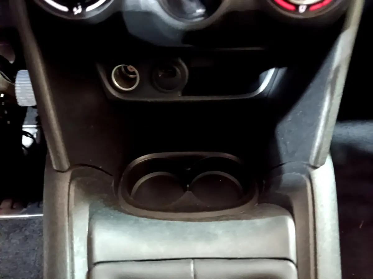 Gros plan sur la console centrale noire de la Peugeot 208 Puretech 82 Signature, avec porte-gobelets et prise 12V visible.