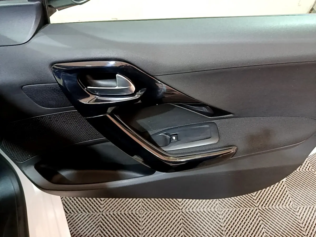 Gros plan sur la porte intérieure côté conducteur d'une Peugeot 208 blanche, avec poignée noire brillante et commande de vitre.
