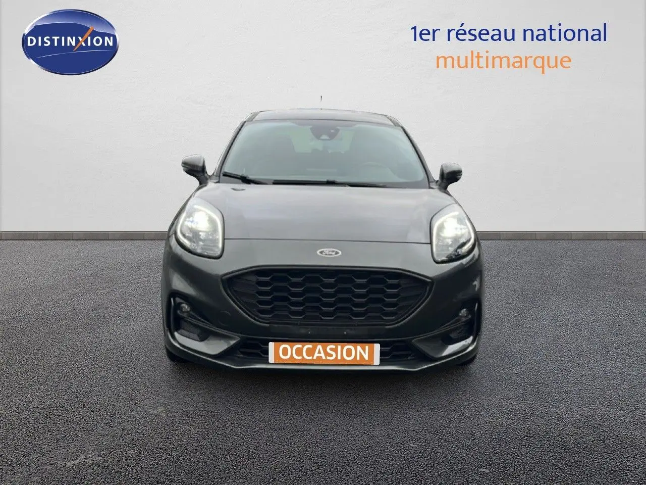 Vue frontale d'un Ford Puma gris magnetic metal 2024 avec calandre noire et phares allumés sur fond neutre.