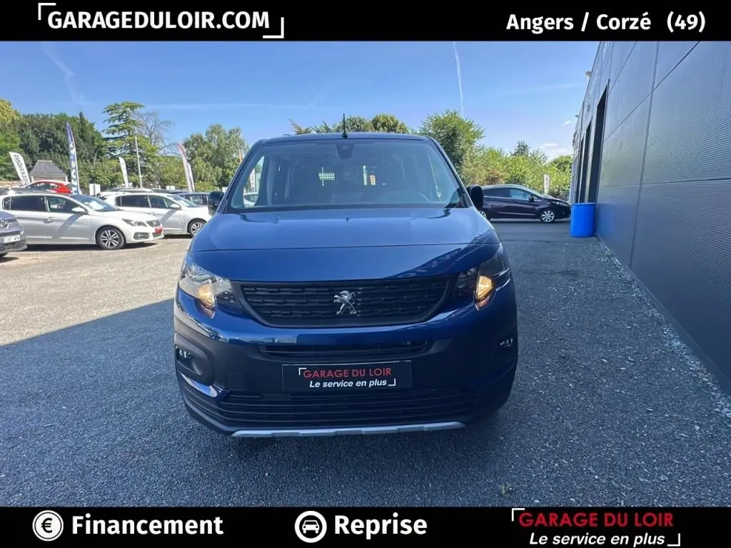 Vue avant frontale du Peugeot Rifter bleu 2024 avec calandre noire et phares allumés sur parking extérieur.