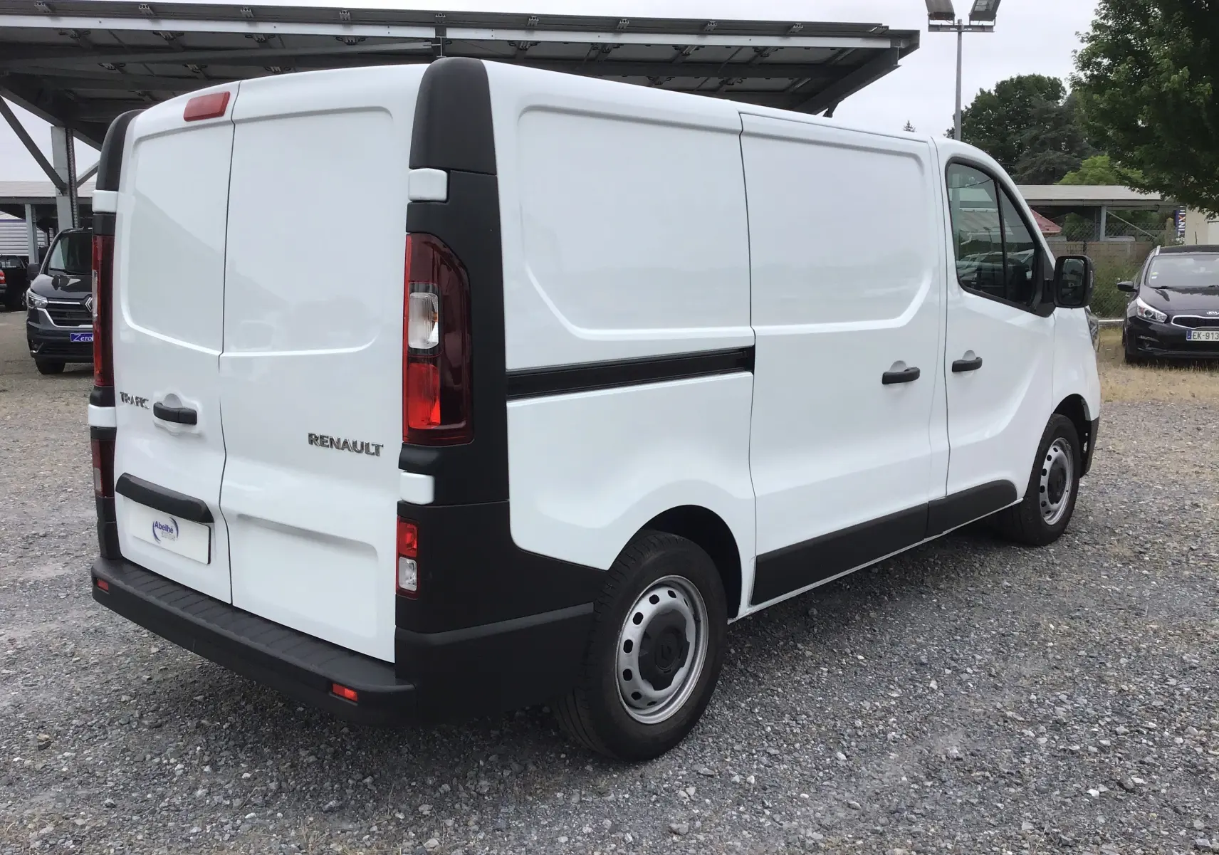 Vue 3/4 arrière droite d’un fourgon Renault Trafic blanc, avec portes arrière fermées et protections noires contrastantes.