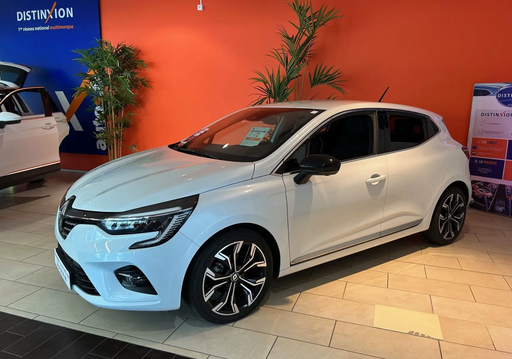Renault Clio V blanc nacré métal vue 3/4 avant droit en showroom avec jantes alliage noires et chromées.
