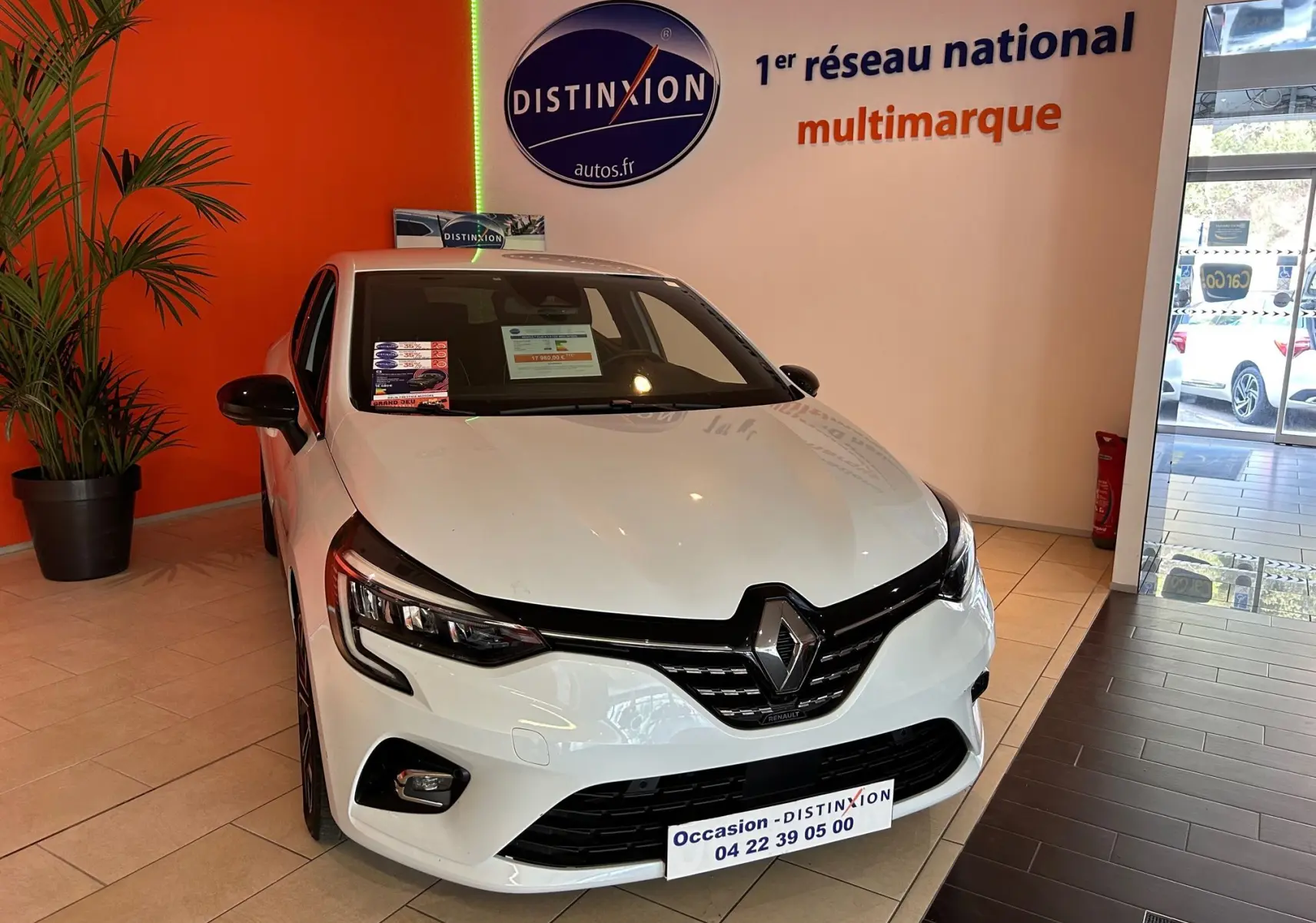 Renault Clio V blanc nacré métal vue 3/4 avant droit en showroom avec logo Distinxion en arrière-plan.