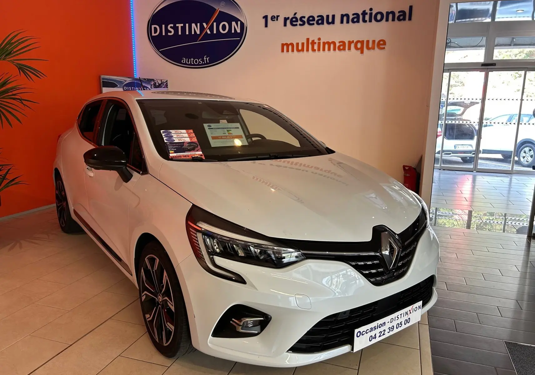Renault Clio V blanc nacré métal vue 3/4 avant droit en showroom avec jantes noires et stickers vitres.
