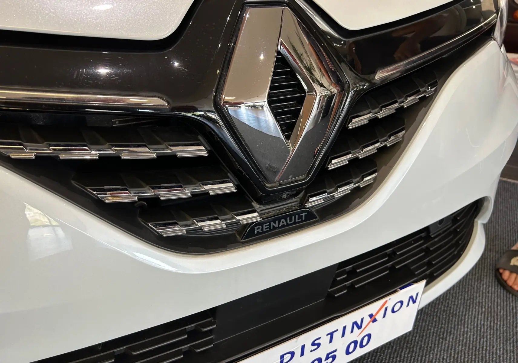 Gros plan sur la calandre avant blanche nacrée d'une Renault Clio V avec logo chrome et plaque du concessionnaire visible.