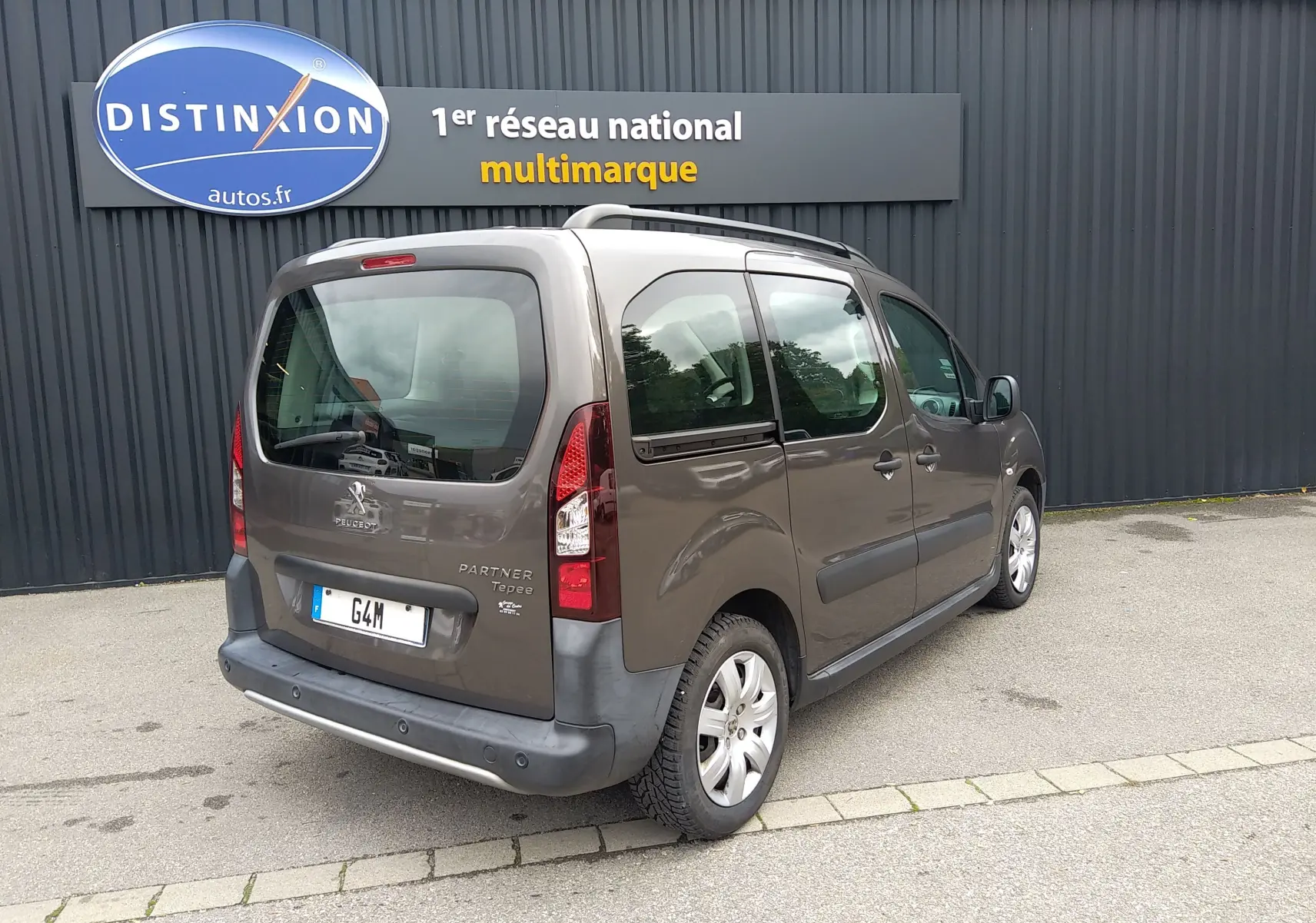 Vue 3/4 arrière droite d'un Peugeot Partner Tepee brun 2017 stationné devant un mur gris avec enseigne Distinxion.