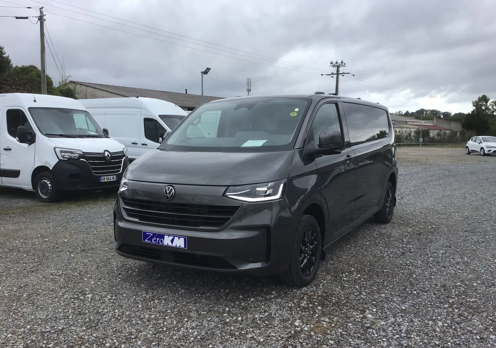Vue 3/4 avant d'un Volkswagen Transporter T7 gris anthracite avec projecteurs LED Matrix et jantes noires sur parking gravel.