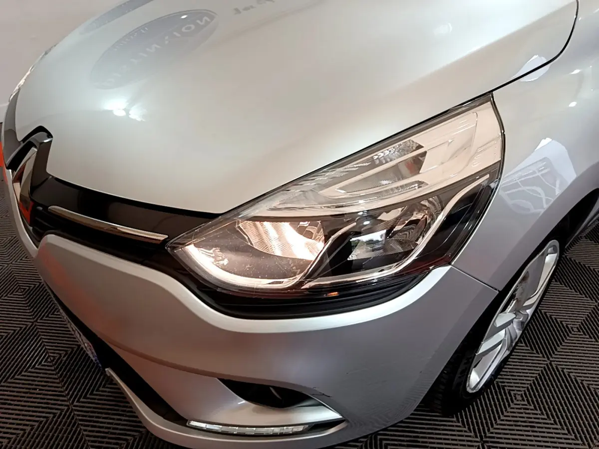 Gros plan sur l'avant droit d'une Renault Clio Business TCe 90 gris clair, mettant en valeur le phare allumé et la calandre noire.