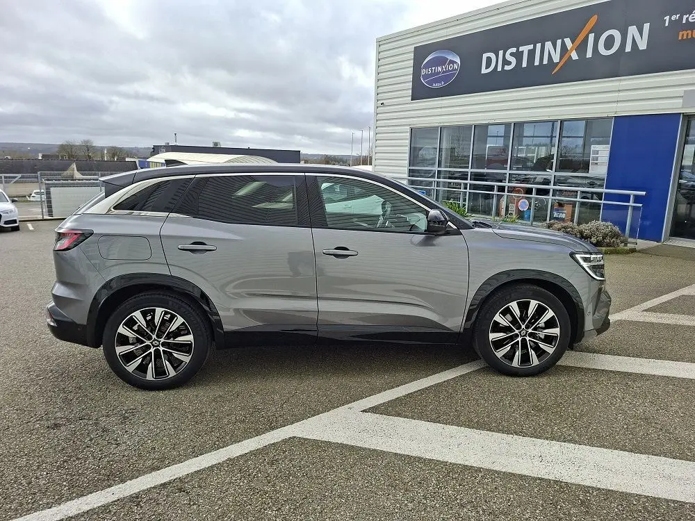 Profil droit d’un Renault Austral E-Tech Full Hybrid gris schiste, avec jantes alliage et vitres teintées, stationné devant un bâtiment.