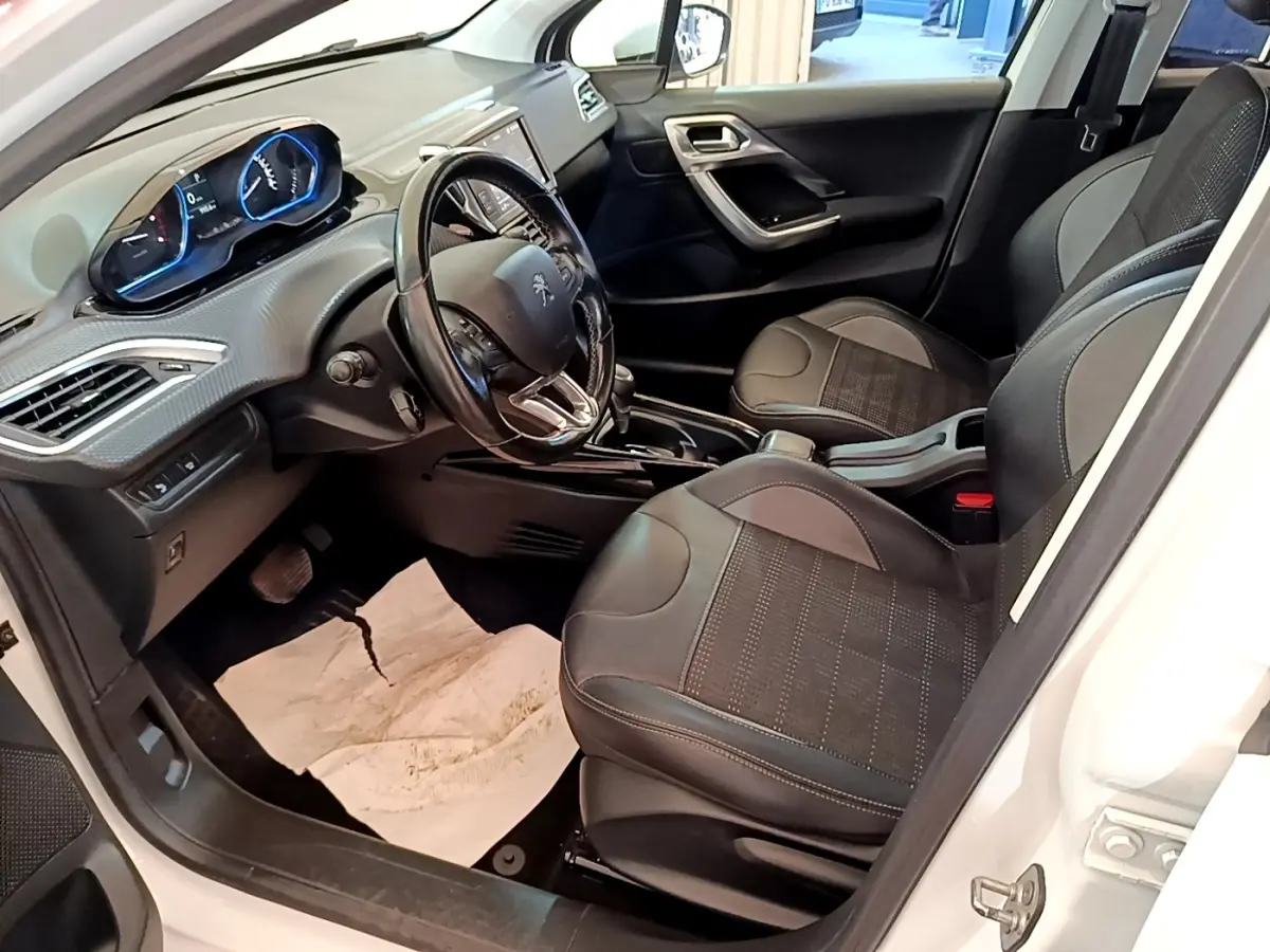 Vue intérieure côté conducteur de la Peugeot 2008 blanche, montrant sièges noirs et tableau de bord avec volant cuir.