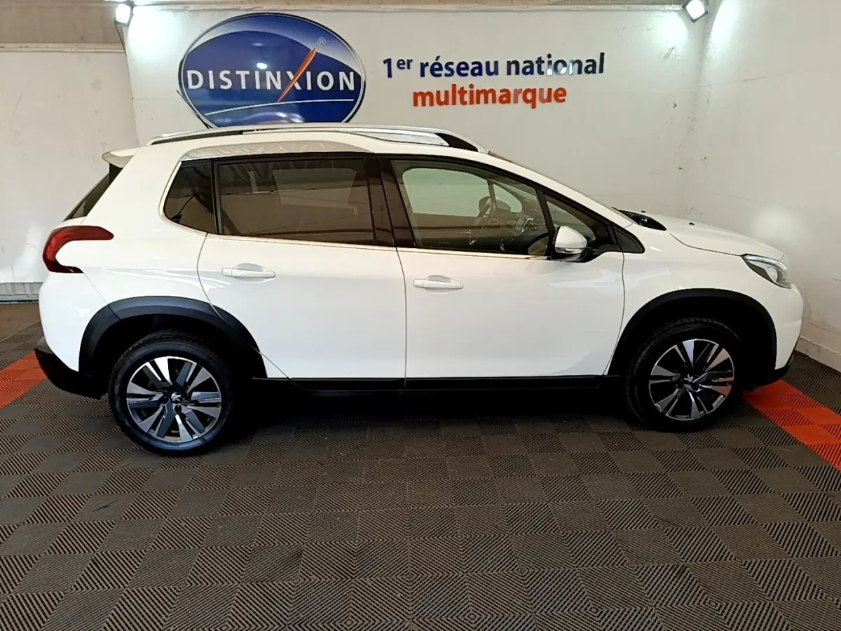 Vue de profil côté gauche d’un Peugeot 2008 blanc avec jantes alliage et barres de toit chromées en intérieur showroom.