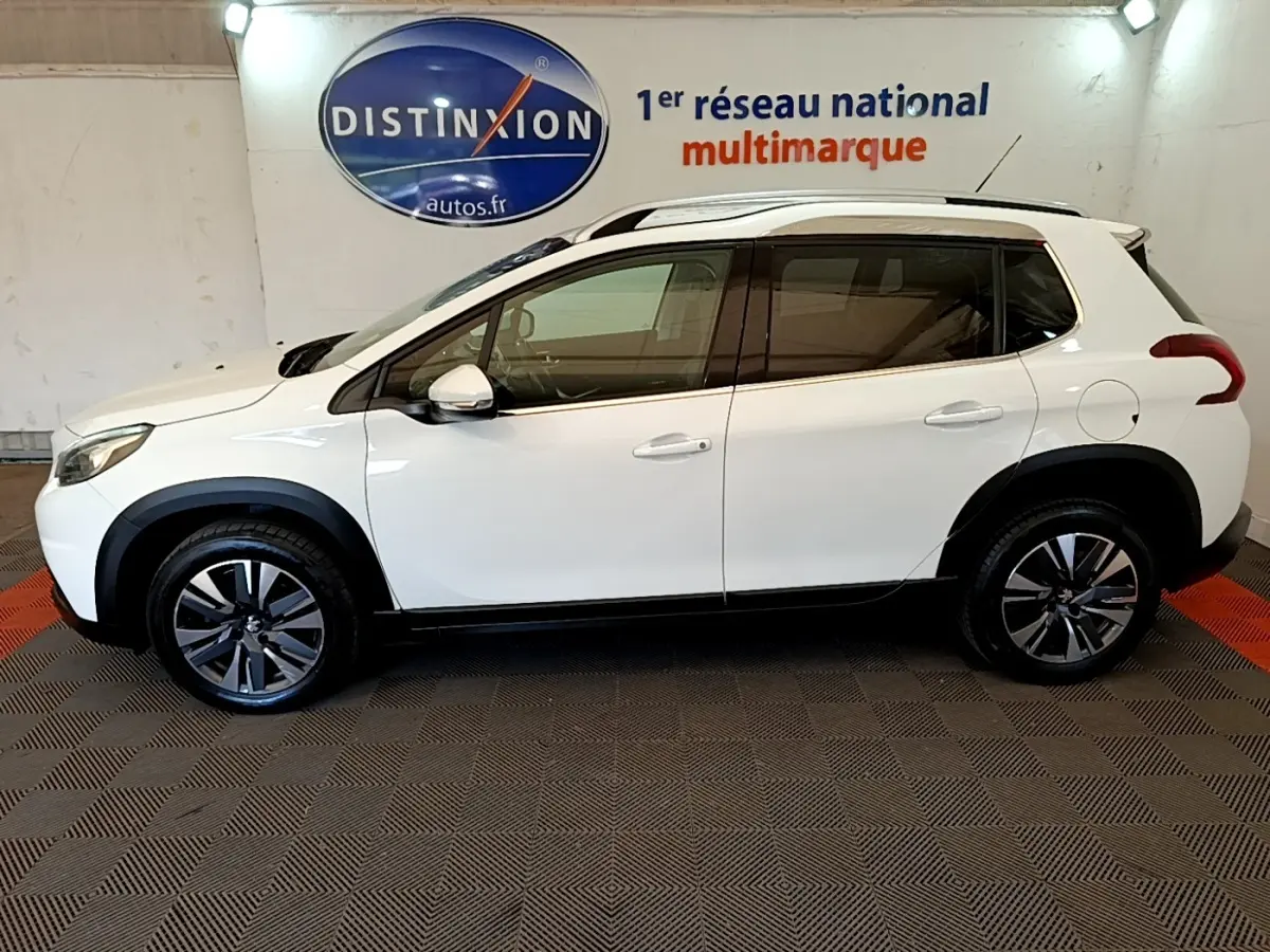 Vue latérale droite d'un Peugeot 2008 blanc avec jantes alliage et barres de toit chromées en intérieur showroom.
