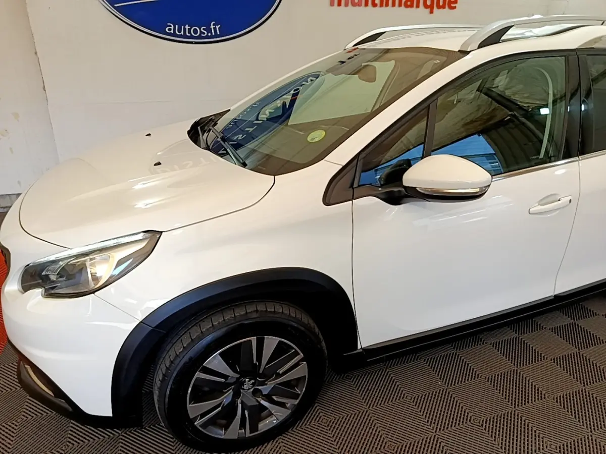 Vue 3/4 avant gauche d’un Peugeot 2008 blanc avec jantes alliage noires et barres de toit chromées.
