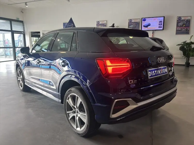 Audi Q2 30 TFSI S-Line bleu Navarre métal vu en 3/4 arrière droit avec feux arrière allumés en showroom.