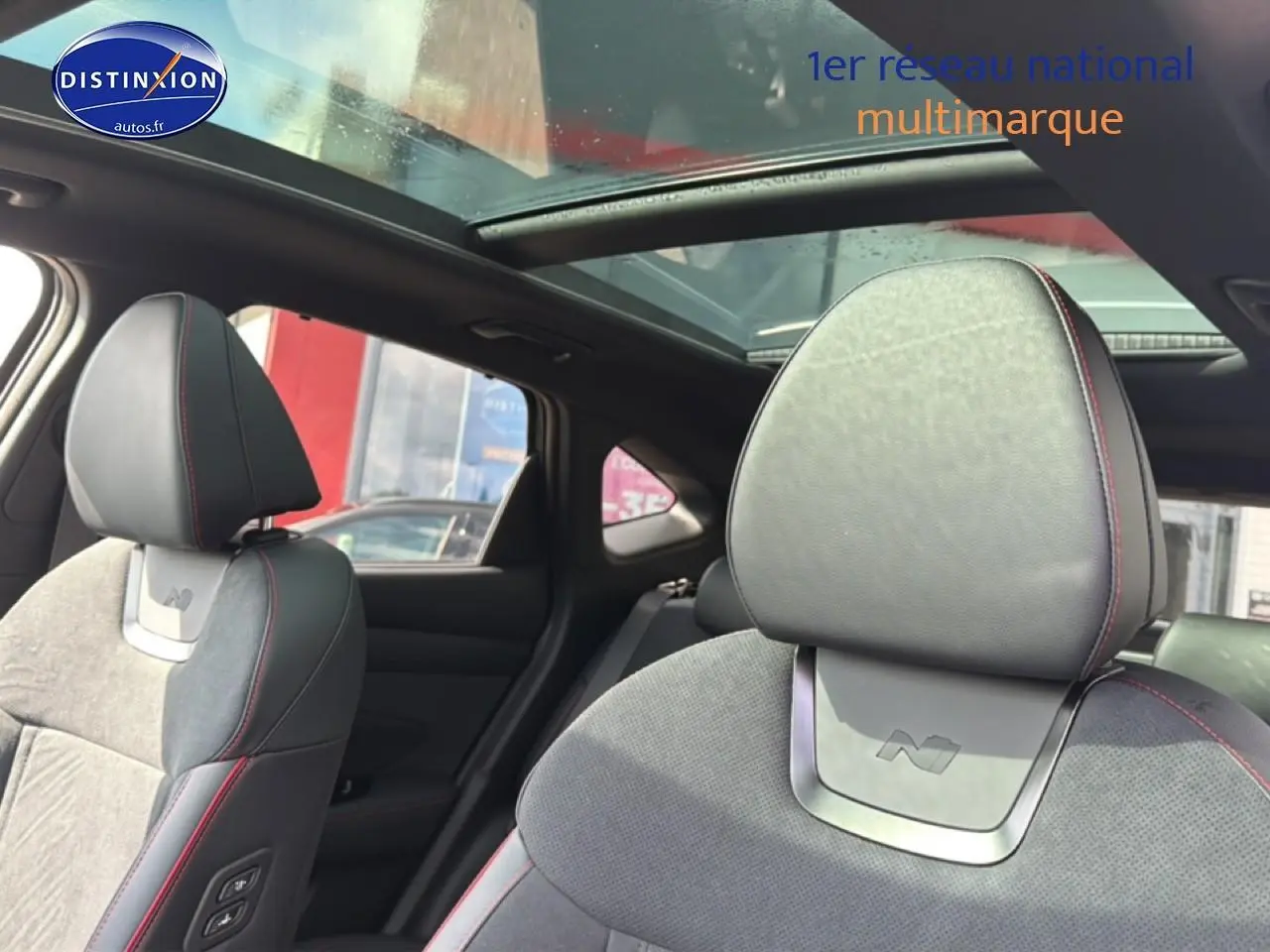Détail intérieur montrant les sièges avant en cuir noir et alcantara avec surpiqûres rouges et toit panoramique en verre.