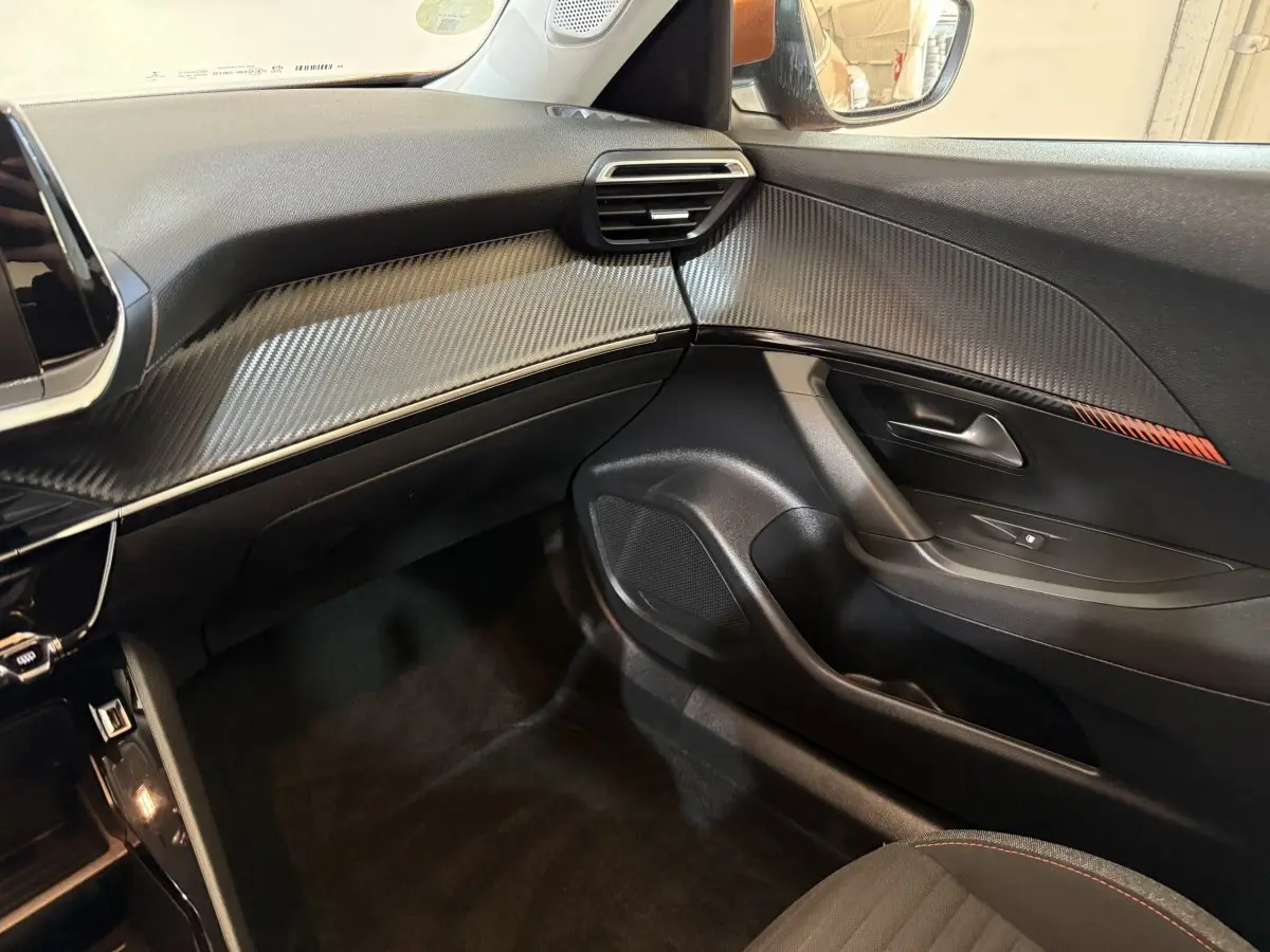 Vue intérieure côté passager du tableau de bord et porte avec insert décoratif effet carbone et surpiqûres orange.