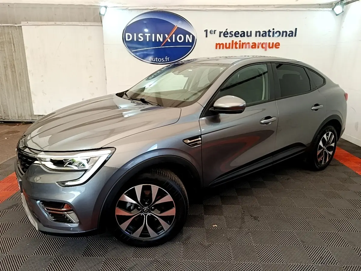Vue 3/4 avant droit d’un Renault Arkana gris clair Evolution E-Tech full hybrid 145 en intérieur showroom.