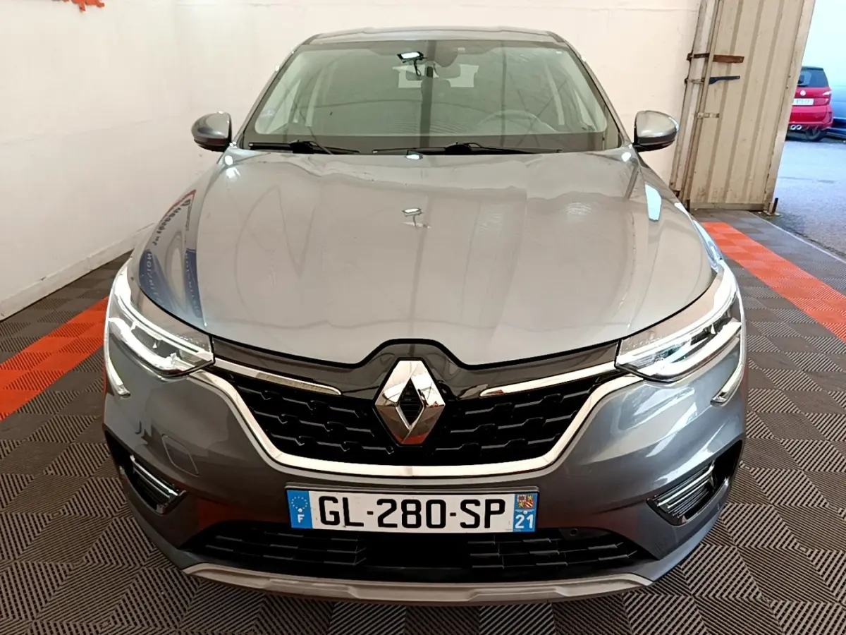 Vue frontale d'un Renault Arkana gris clair 2023 avec calandre noire et phares LED allumés dans un garage.