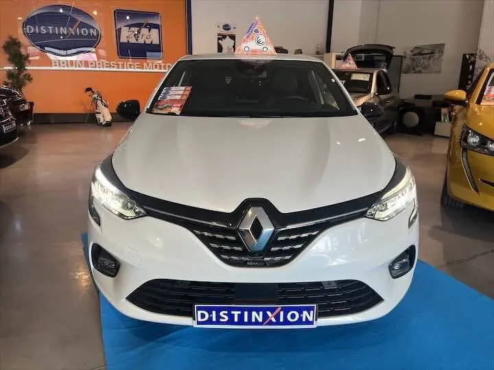 Vue avant d'une Renault Clio V blanc nacré métal avec phares allumés et plaque Distinxion en concession