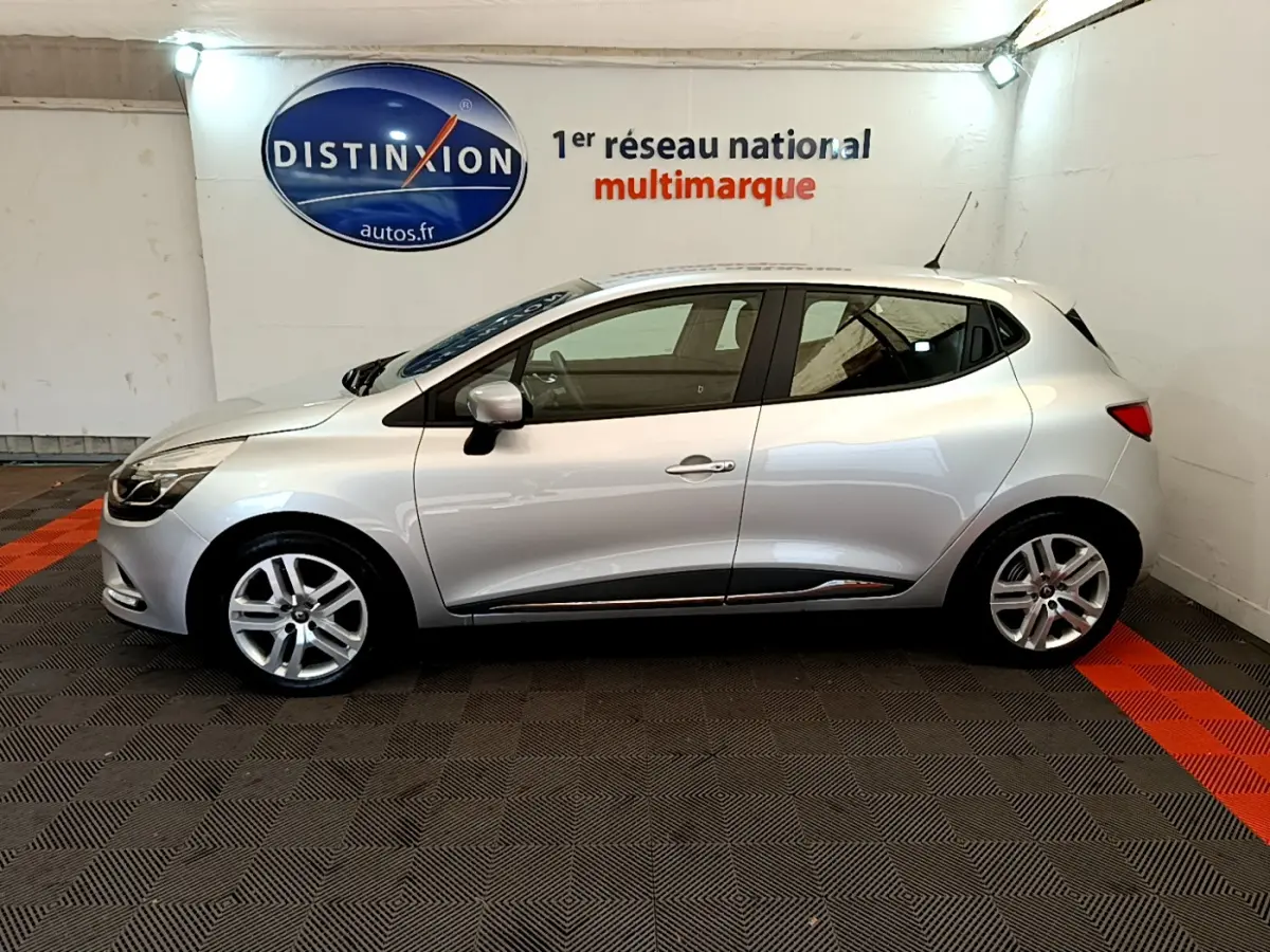 Profil côté gauche d'une Renault Clio Business TCe 90 gris clair, en intérieur sur sol noir et rouge, avec logo Distinxion en arrière-plan.