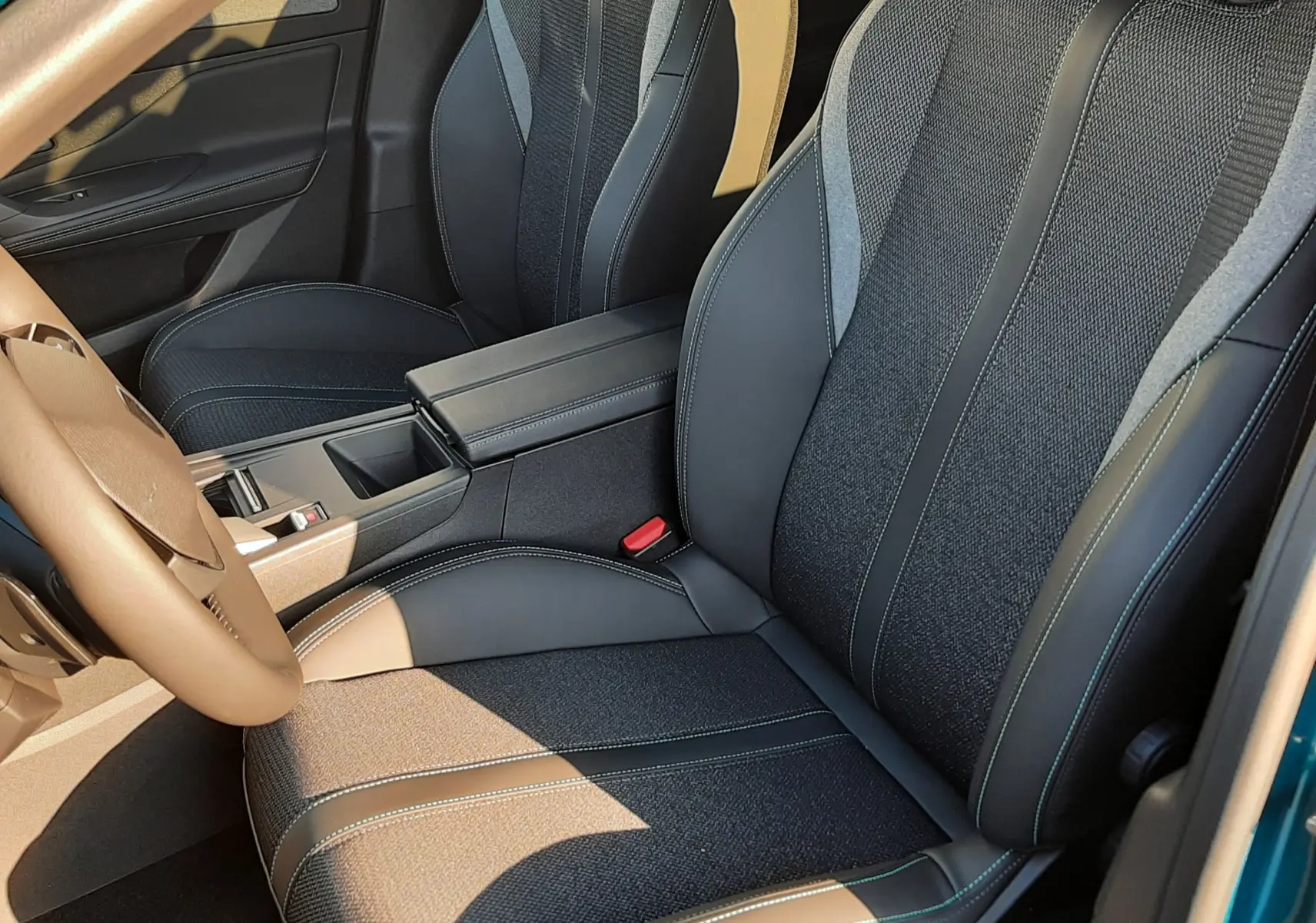 Vue intérieure côté conducteur de la Peugeot 308 2025, sièges noirs en tissu et cuir avec surpiqûres vertes, volant beige visible.
