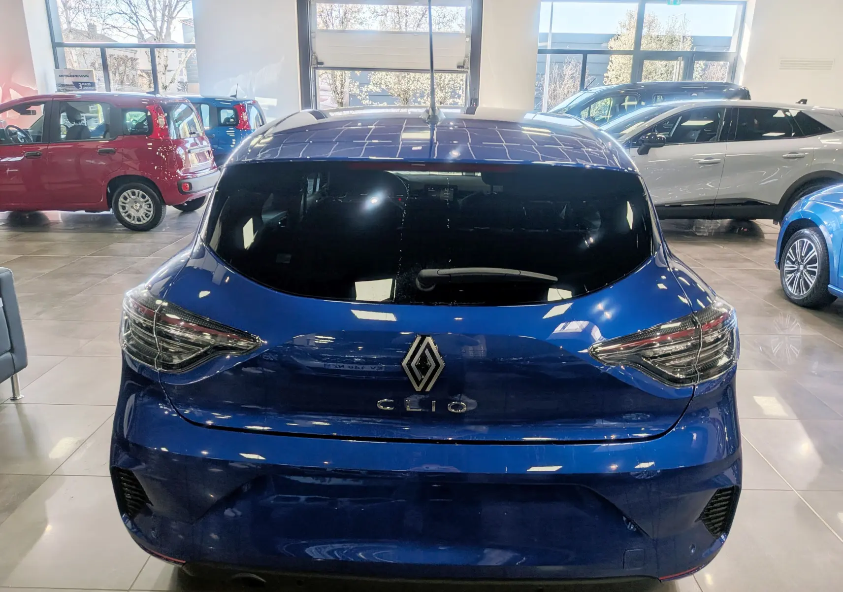 Vue arrière d'une Renault Clio V Bleu Iron dans un showroom, avec feux arrière LED et vitre arrière teintée.