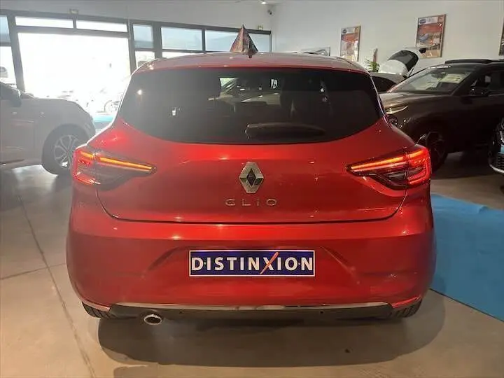 Vue arrière d'une Renault Clio V rouge flamme métal avec feux allumés et plaque Distinxion en concession.
