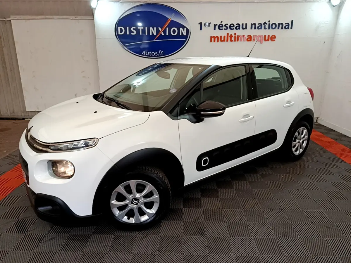 Citroën C3 blanche vue 3/4 avant droit, avec protections latérales noires et jantes alliage en intérieur showroom.