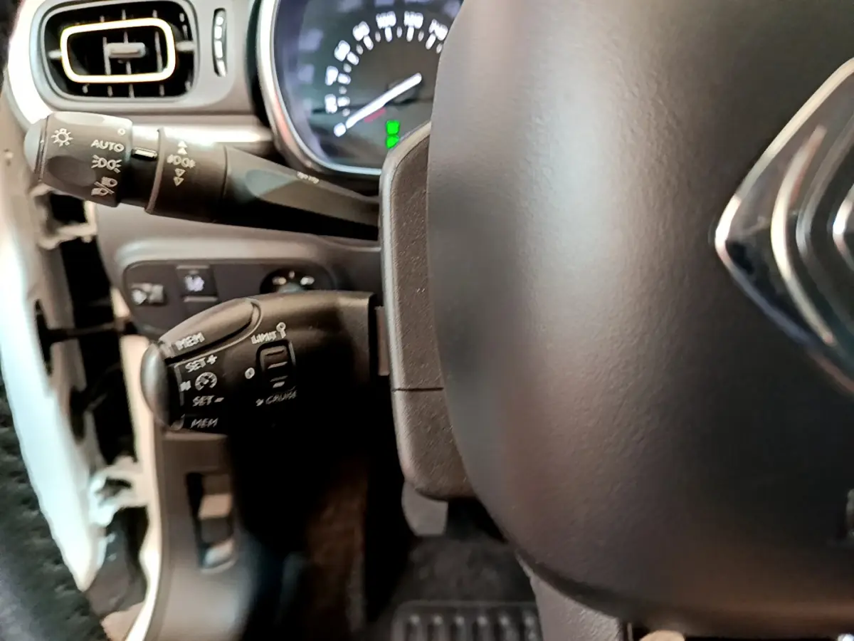 Gros plan sur le volant cuir et les commandes de régulateur de vitesse à gauche du tableau de bord d'une Citroën C3 blanche.