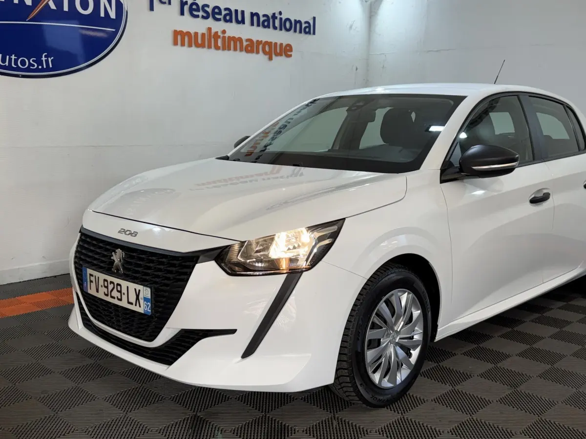Peugeot 208 blanche vue 3/4 avant droit, phare allumé et calandre noire distinctive sur fond intérieur showroom.