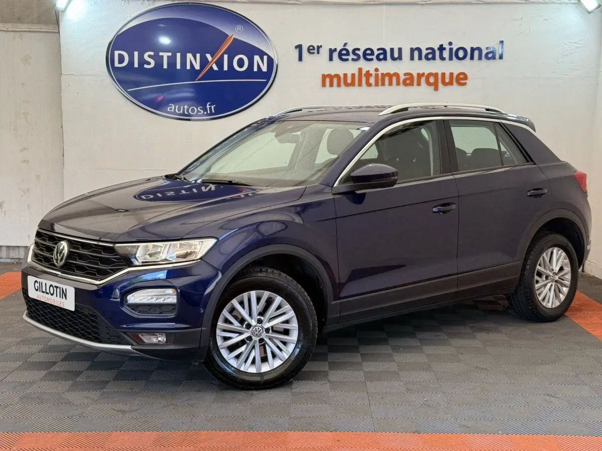 Volkswagen T-Roc bleu en 3/4 avant droit, avec jantes alliage et barres de toit noires visibles.